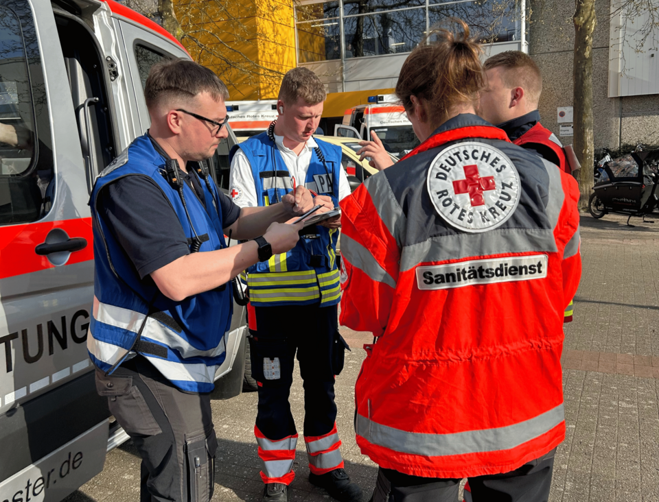 Vier Personen in Einsatzkleidung stehen zusammen und besprechen sich. Im Hintergrund sind Einsatzfahrzeuge zu sehen.