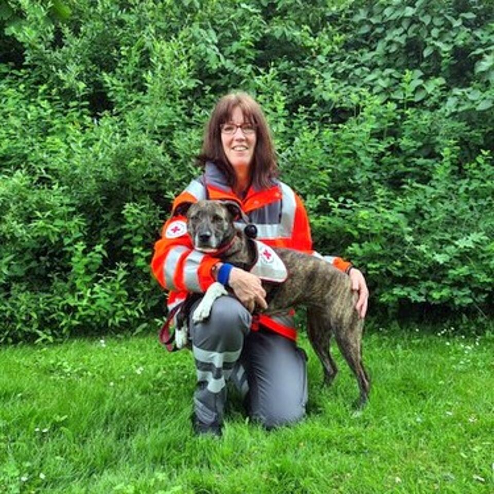 Eine Person in Einsatzkleidung kniet neben einem Hund. Im Hintergrund befindet sich grüne Vegetation.