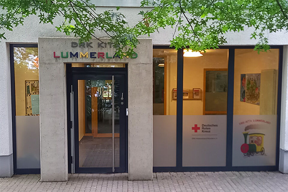 Eingang eines Gebäudes mit dem Schild ‚DRK KITA LUMMERLAND‘ über der Tür. Der Schriftzug ‚LUMMERLAND‘ ist in bunten Buchstaben gestaltet. Links und rechts der Tür befinden sich große Glasfenster mit Dekorationen und Plakaten. Auf einem Fenster ist das Logo des Deutschen Roten Kreuzes sowie eine Illustration eines Zuges mit dem Schriftzug ‚DRK-KITA LUMMERLAND‘ zu sehen