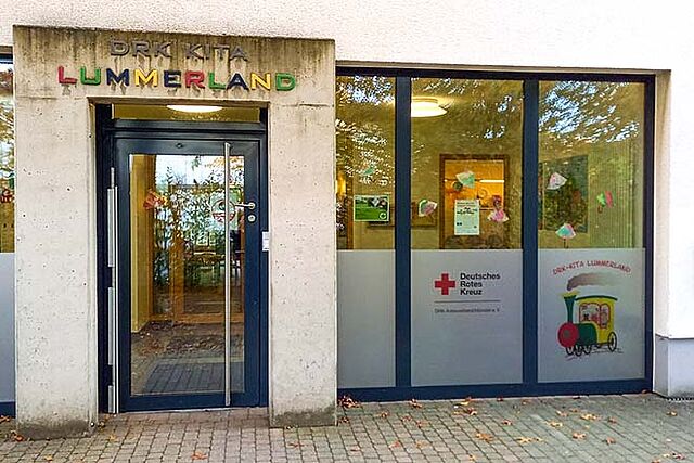 Eingang eines Gebäudes mit dem Schild ‚DRK KITA LUMMERLAND‘ über der Tür. Der Schriftzug ‚LUMMERLAND‘ ist in bunten Buchstaben gestaltet. Links und rechts der Tür befinden sich große Glasfenster mit Dekorationen und Plakaten. Auf einem Fenster ist das Logo des Deutschen Roten Kreuzes sowie eine Illustration eines Zuges mit dem Schriftzug ‚DRK-KITA LUMMERLAND‘ zu sehen