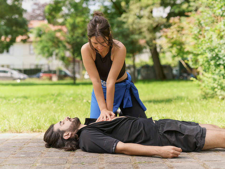 Eine Frau führt im Freien eine Wiederbelebung durch. Sie kniet auf dem Boden und drückt mit beiden Händen auf den Brustkorb eines Mannes, der auf dem Rücken liegt. Die Umgebung besteht aus Rasen, Bäumen und einigen Gebäuden im Hintergrund.