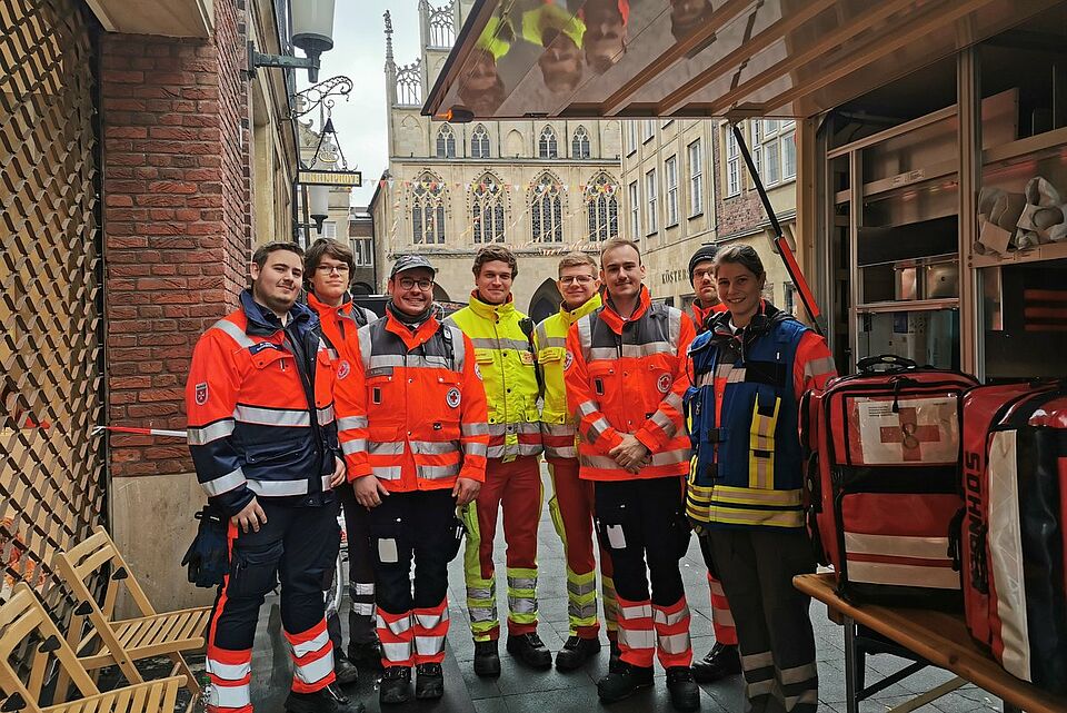 Rosenmontag Acht Personen in Einsatzkleidung verschienender Hilfsorganisation stehen neben einem Gerätewagen. Im Hintergrund ist das historische Rathaus zu sehen