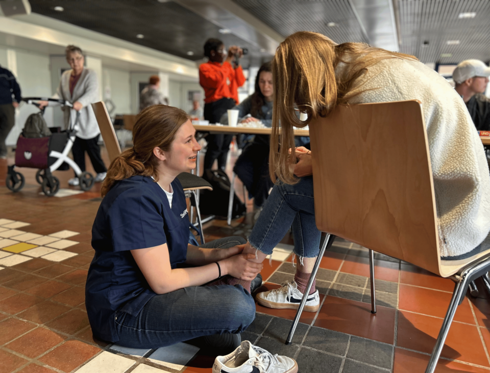 Eine Person sitzt mit gesenktem Kopf auf einem Stuhl. Eine Person sitzt vor ihr auf dem Boden, hält ihr Bein und spricht mir ihr. Im Hintergrund stehen Tische, an denen weitere Personen sitzen.