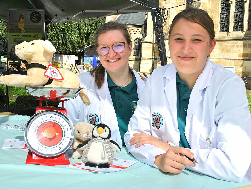 Zwei Personen sitzen an einem Tisch auf dem eine manuelle Waage und mehrere Kuscheltiere stehen