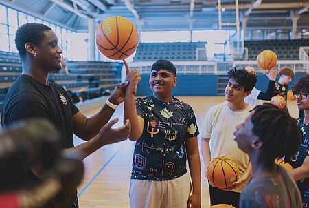 In einer Sporthalle stehen mehrere Jugendliche mit Basketbällen um einen Mann herum, der ihnen einen Trick mit dem Ball zeigt
