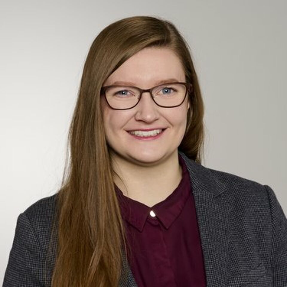 Frau mit braunen Haaren, Brille, roter Bluse und grauem Blazer