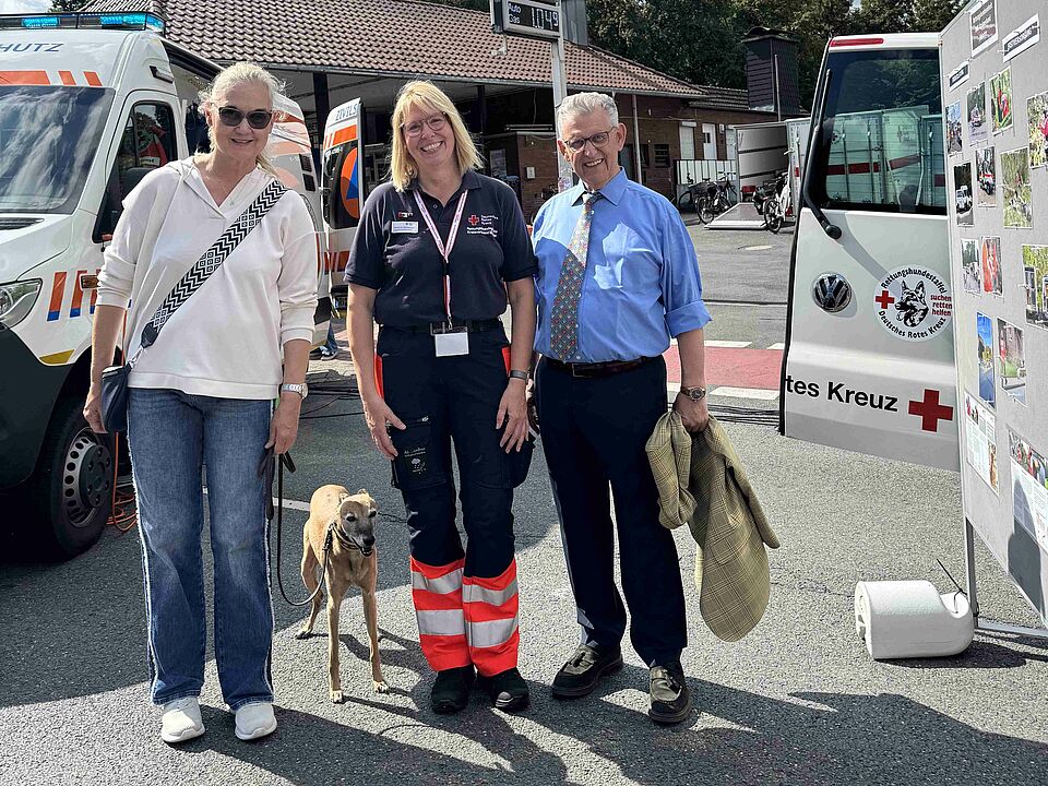 Drei Personen stehen in einer Reihe mit einem Hund, im Hintergrund ist ein Krankenwagen zu sehen