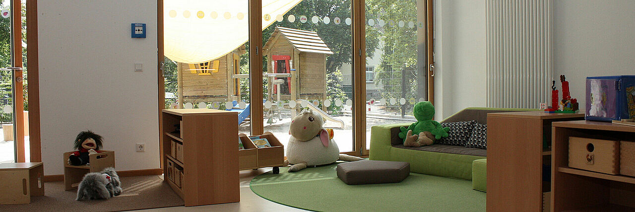 Meerwiese Gruppenraum mit Kindermöbeln und Spielzeug, große Fensterfront wo Außenbereich mit Spielgeräten zu sehen sind