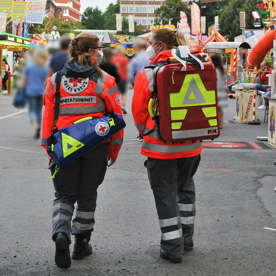 Zwei Personen in Einsatzkleidung gehen von der Kamera weg. Auf den Jacken steht „DEUTSCHES ROTES KREUZ Rettungsdienst“. Beide tragen medizinische Taschen: eine blaue mit gelbem Dreieck und rotem Kreuz, die andere rot mit ähnlichen Symbolen. Die Szene spielt sich im Freien ab, im Hintergrund sind Fahrgeschäfte und Personen zu sehen.