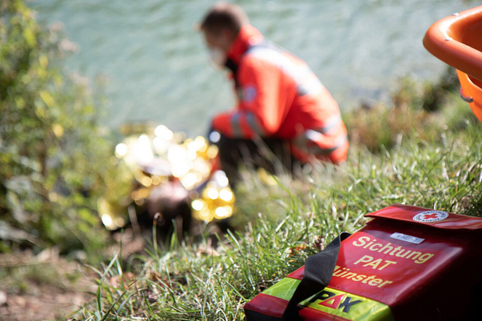 Am Ufer eines Gewässers liegt eine Person auf dem Boden, zugedeckt mit einer silbernen Rettungsdecke. Eine Einsatzkraft in Einsatzkleidung leistet Hilfe. Im Vordergrund steht eine rote Notfalltasche mit gelben und grünen Markierungen. Auf der Tasche steht: „Sichtung PAT Münster"