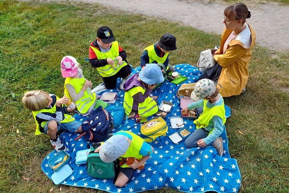Sieben Kinder in Warnwesten sitzen mit einer erwachsenen Person auf einer Picknickdecke auf einer Wiese.