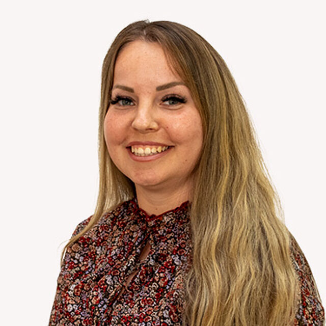 Frau mit langen blonden Haaren und gemusterter Bluse vor weißem Hintergrund