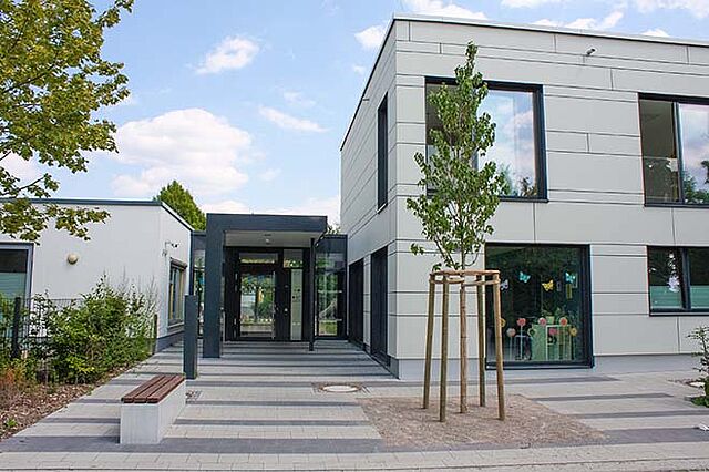 Ein modernes Gebäude mit flachem Dach, großen Fenstern und einer hellen Fassade mit horizontalen Linien. Vor dem Gebäude befindet sich eine gepflasterte Fläche mit Bäumen und Sträuchern. Im Vordergrund steht ein junger Baum, der von Holzpfählen gestützt wird. Der Eingang mit Glastüren und einem Vordach ist gut sichtbar
