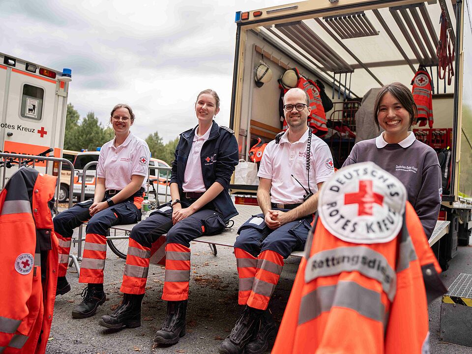 Vier Personen in Einsatzkleidung sitzen auf der Ladefläche von einem LKW, im Hintergrund sind DRK-Fahrzeuge zu sehen