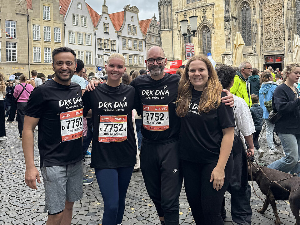 Vier Personen stehen Arm in Arm in schwarzen T-Shirts mit DRK-Aufschrift. Im Hintergrund sind Menschen und historische Häuser zu sehen.