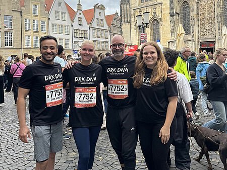 Vier Personen stehen Arm in Arm in schwarzen T-Shirts mit DRK-Aufschrift. Im Hintergrund sind Menschen und historische Häuser zu sehen.