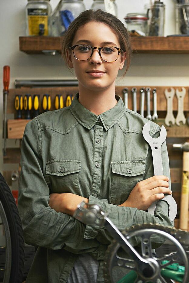 Eine Person steht in einer Werkstatt oder Garage und hält einen großen Schraubenschlüssel in der Hand. Im Hintergrund hängt an einer Lochwand eine Vielzahl von Werkzeugen, darunter Schraubenschlüssel, Schraubendreher und Zangen. Darüber befinden sich Regale mit Gläsern und Behältern. Im Vordergrund ist ein Teil eines Fahrrads zu sehen, insbesondere das Rad und das Tretlager.