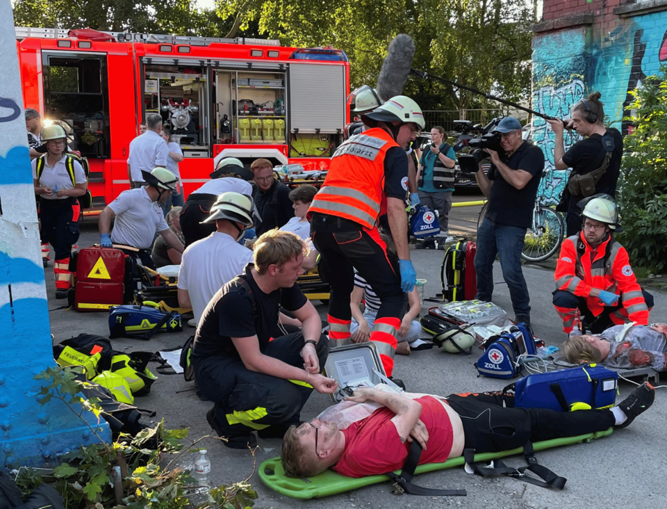 Mehrere Verletztende liegen auf Tragen auf dem Boden und werden von Einsatzkräften versorgt. Im Hintergrund ist ein Feuerwehreinsatzfahrzeug zu sehen.