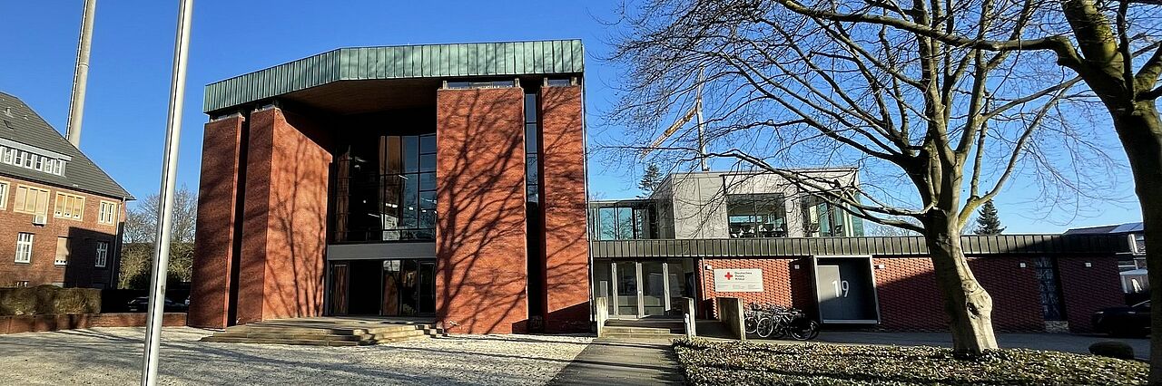 Ein Gebäude aus roten Ziegelsteinen steht im Zentrum des Bildes. Es ist von Bäumen und Pflanzen umgeben. Links im Bild ragen Fahnenmast ohne Flaggen empor. Der Himmel ist klar und blau.