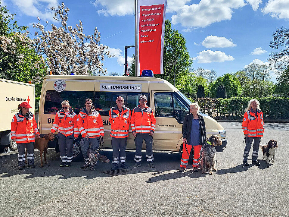 Mantrailing Prüfung Sieben Personen in Einsatzkleidung und jeweils einem Hund an der Leine stehen vor einem Einsatzfahrzeug mit der Beschriftung Rettungshunde. Im Hintergrund ist eine DRK-Fahne zu sehen.