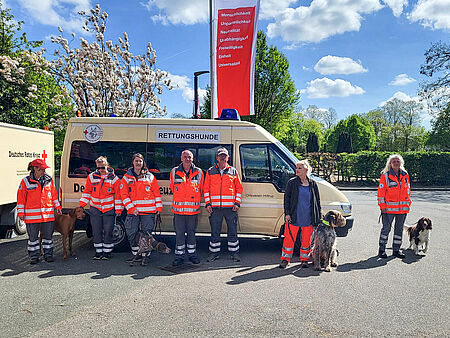 Sieben Personen in Einsatzkleidung und jeweils einem Hund an der Leine stehen vor einem Einsatzfahrzeug mit der Beschriftung Rettungshunde. Im Hintergrund ist eine DRK-Fahne zu sehen.