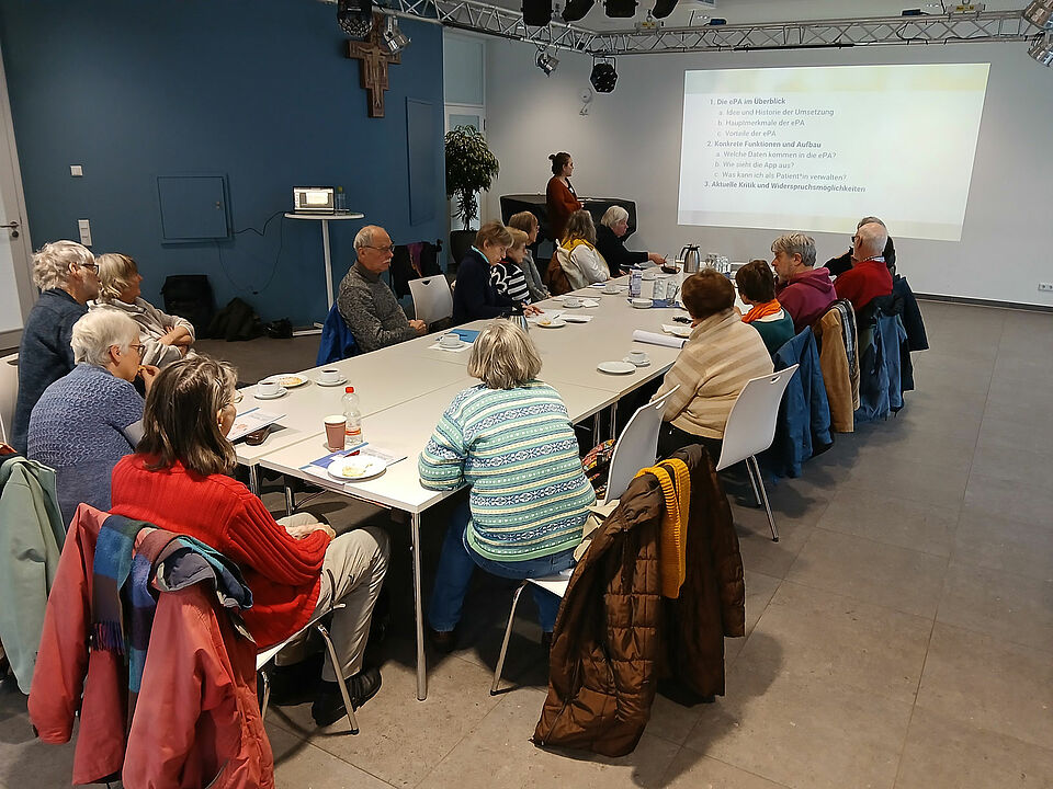 Eine Gruppe von älteren Menschen sitzt um einen Tisch herum und schaut auf eine Präsentation an der Wand, neben der eine Referentin steht.
