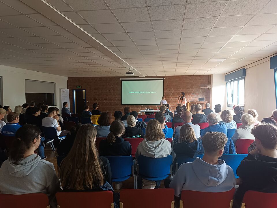 Publikum Ein Raum mit vollem Publikum, vorne stehen zwei Personen auf einer Art Bühne neben einer Leinwand