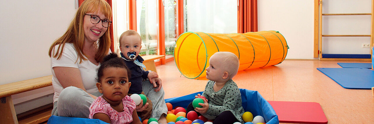 Wolkenburg Eine Person mit drei kleinen Kindern sitzt auf dem Boden in einem Ballebad, im Hintergrund eine Sprossenwand