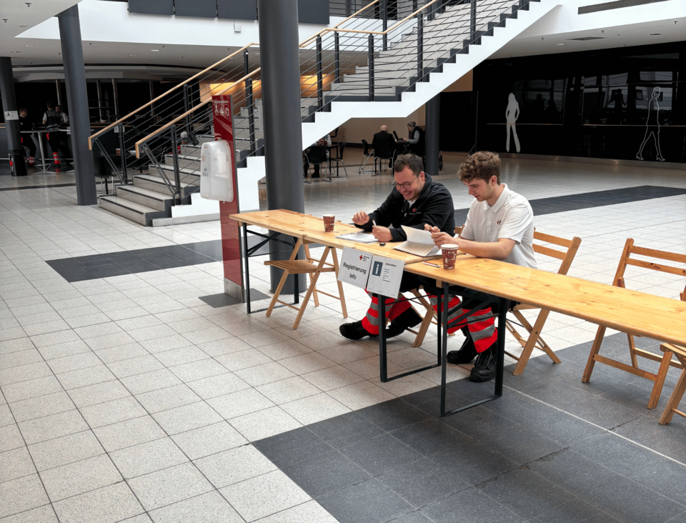 Zwei Personen in Einsatzkleidung sitzen an einem langen Holztisch in einem Eingangsbereich