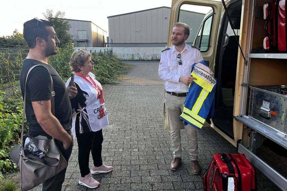 Besuch aus Chile Eine Person steht vor einem offenen Geräteeinsatzwagen und zeigt zwei weiteren Personen eine blaue Weste mit der Aufschrift "Patientenablage"