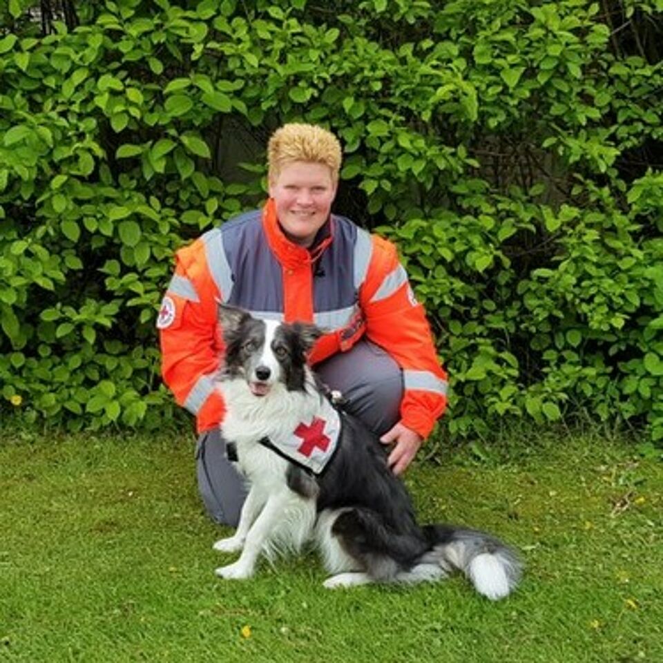 Eine Person in Einsatzkleidung kniet neben einem Hund. Im Hintergrund befindet sich grüne Vegetation.