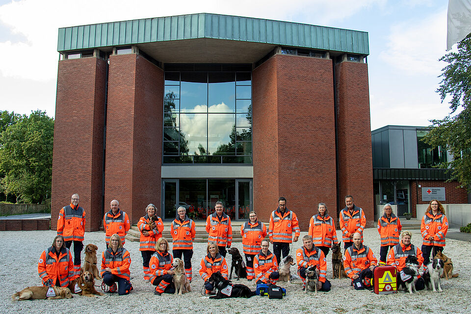 Eine Gruppe von Personen in Einsatzkleidung steht vor einem modernen Backsteingebäude mit großen Glasfenstern. Mehrere Hunde befinden sich bei der Gruppe, einige sitzen, andere stehen.