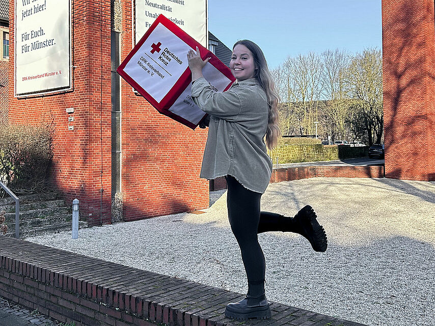 Eine Person balanciert auf einem Bein auf einer niedrigen Mauer und hält eine große rot-weiße Box mit dem Logo des Deutschen Roten Kreuzes hoch. Im Hintergrund ist ein rotes Backsteingebäude mit zwei großen Schildern zu sehen: „Jetzt hier! Für Euch. Für Münster.“ und „Neutralität Unabhängigkeit Menschlichkeit“.