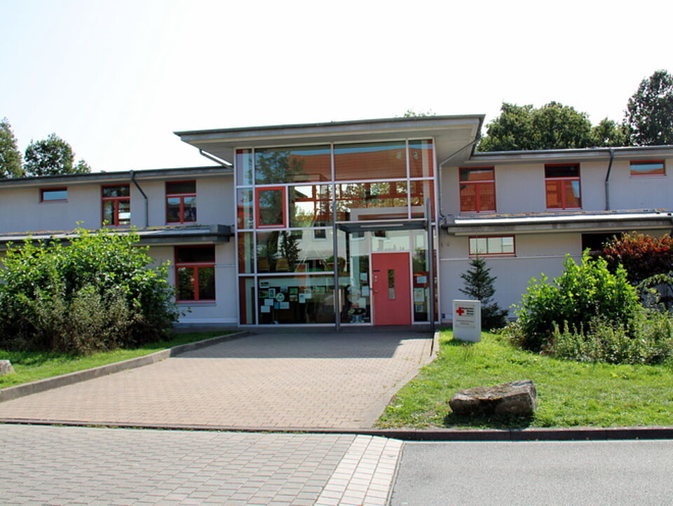 Wolkenburg Ein modernes, zweistöckiges Gebäude mit großen Fenstern und einer roten Eingangstür. Ein gepflasterter Weg führt durch grüne Büsche und Bäume zum Eingang.