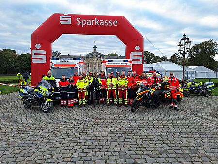 Eine Gruppe von Einsatzkräften vor einem Bogen mit Sparkassenlogo. An den Rändern steht jeweils ein Motorrad. Im Hintergrund sind Zelte und ein historisches Schloss zu sehen.