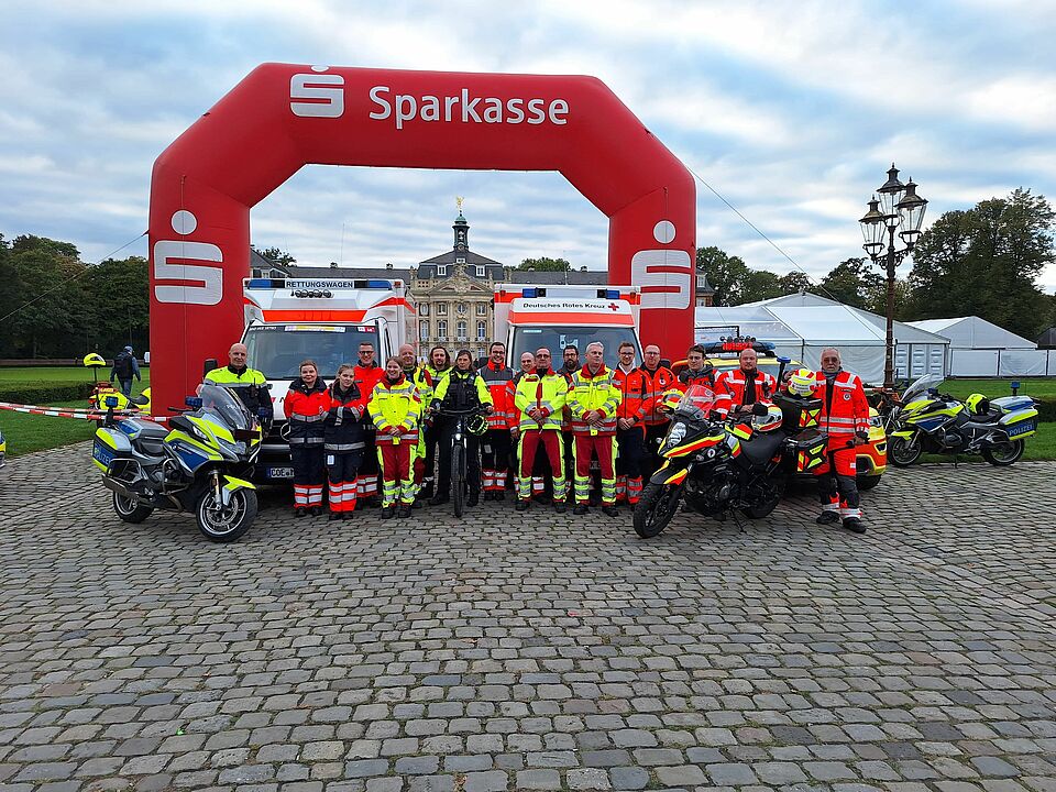 Giro2 Eine Gruppe von Einsatzkräften vor einem Bogen mit Sparkassenlogo. An den Rändern steht jeweils ein Motorrad. Im Hintergrund sind Zelte und ein historisches Schloss zu sehen.