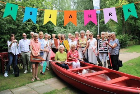 Eine Gruppe von Menschen steht auf einer Wiese. Vor ihnen ist ein Kanu, in dem zwei Personen sitzen. Mehrere halten Paddel in der Hand. Am oberen Rand des Bildes wurde digital eine Wimpelkette mit der Aufschrift "10 Jahre" hinzugefügt.
