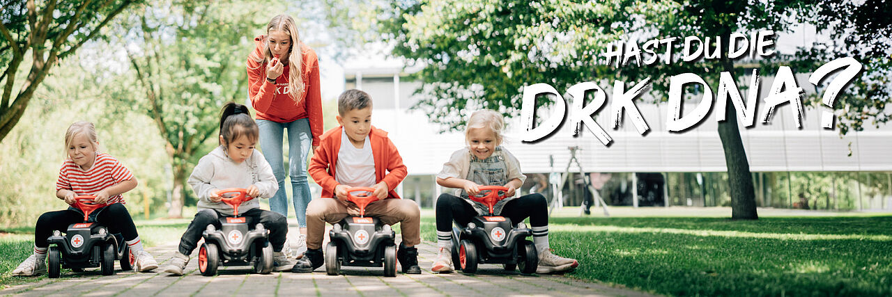Fünf Kinder sitzen auf Spielautos in einer parkähnlichen Umgebung mit Bäumen und Gras. Eine erwachsene Person im roten Shirt steht dahinter. Rechts im Bild steht der Text: „#Hast du die DRK DNA?“