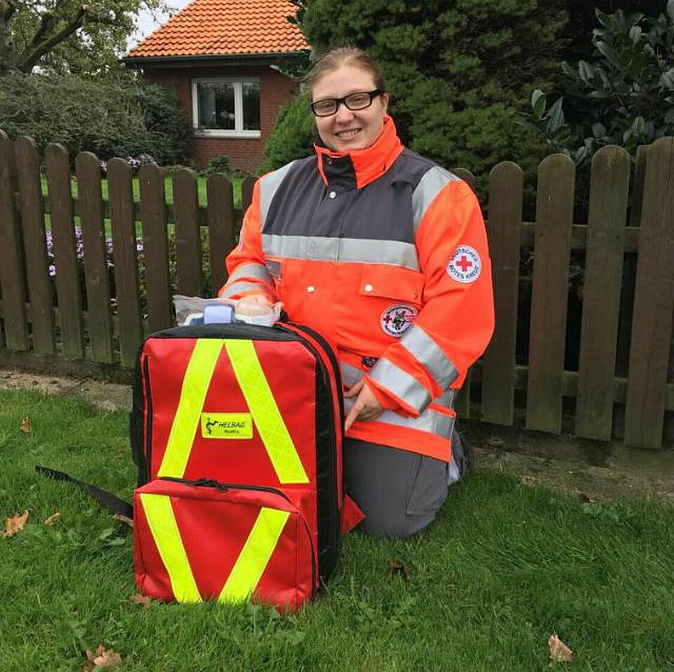 Eine Person in Einsatzkleidung kniet auf einer Wiese neben einem Holzzaun. Vor ihr steht ein großer roter Rucksack mit gelben Reflektorstreifen und der Aufschrift „HELMAG Rettungsdienst“. Auf dem Ärmel der Kleidung ist ein Rotkreuz-Emblem zu sehen.