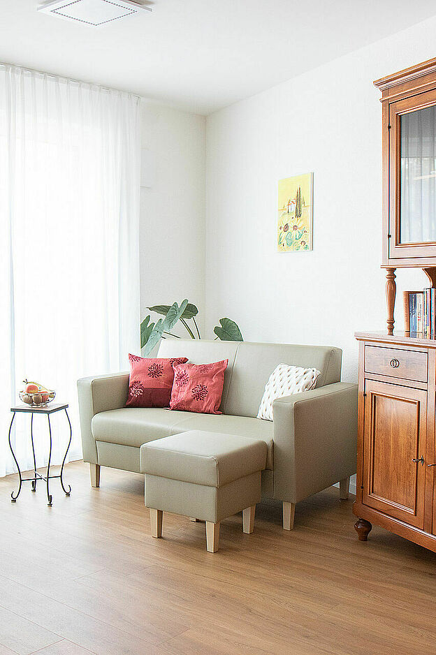 Wohnzimmer mit Holzfußboden und großer Fensterfront mit weißen Vorhängen. Links ein kleiner Tisch mit Dekoration und eine Topfpflanze, daneben ein hoher Holzschrank mit Kerzenständern. Rechts ein L-förmiges beiges Sofa mit roten und weißen Kissen, darüber ein gerahmtes Bild. Daneben ein großer Holzschrank mit Büchern und Ausstellungsstücken hinter Glas