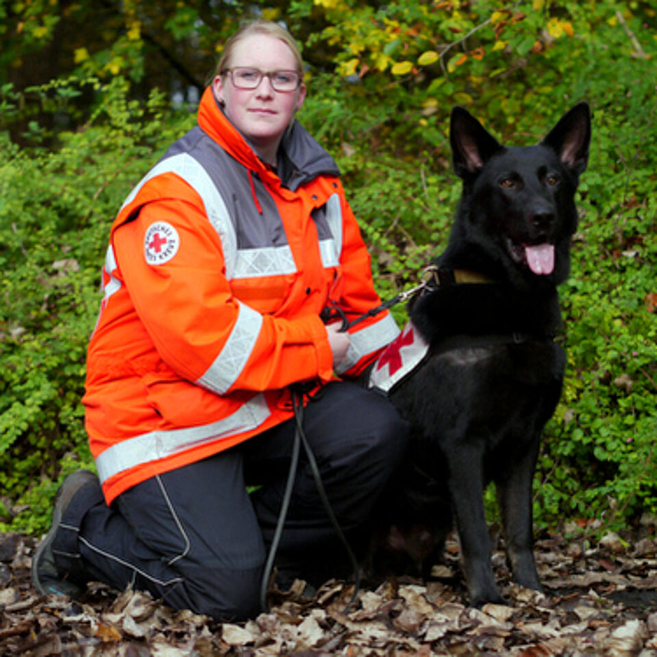 Eine Person in Einsatzkleidung kniet neben einem Hund. Im Hintergrund befindet sich grüne Vegetation.