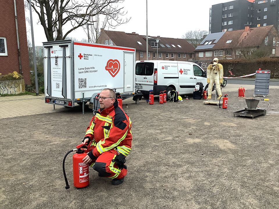 Im Vordergrund hockt eine Person in Einsatzkleidung und einem Feuerlöscher. Im Hintergrund befindet sich ein Auto und Anhänger mit DRK-Beschriftung sowie eine Puppe und weitere Feuerlöscher.