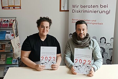 Zwei Personen halten eine Broschüre mit dem Titel "Antidiskriminierungsberatung der Freien Wohlfahrtspflege NRW: Jahresbericht 2022" in der Hand, im Hintergrund ein Aufstellung der Antidiskriminierungsberatungen und Flyerständer