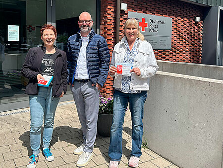 Vor dem Gebäude des Deutschen Roten Kreuzes in Münster stehen drei Personen lächelnd für ein Foto. Zwei Frauen halten die Preise in der Hand