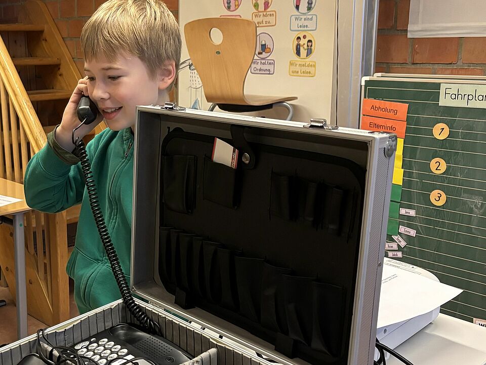 Ein Kind hat einen Telefonhörer am Ohr und steht neben einem Koffer mit einem Telefon und viel Technik. Im Hintergrund befindet sich eine Schultafel mit Post-its.