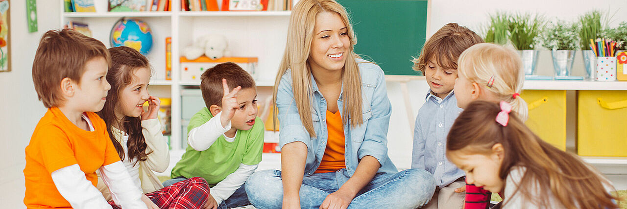 Kita Slider Sechs Kinder sitzen um eine erwachsene Person herum, im Hintergrund Regale mit Spielmaterialien