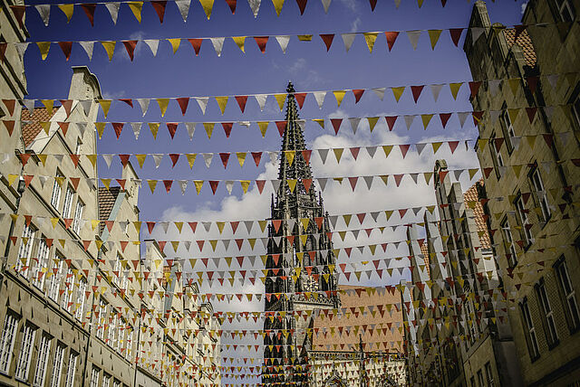 Straßenszene mit bunten Wimpelketten, die zwischen den Gebäuden gespannt sind. Im Hintergrund ist eine Kirche mit spitzem Turm zu sehen