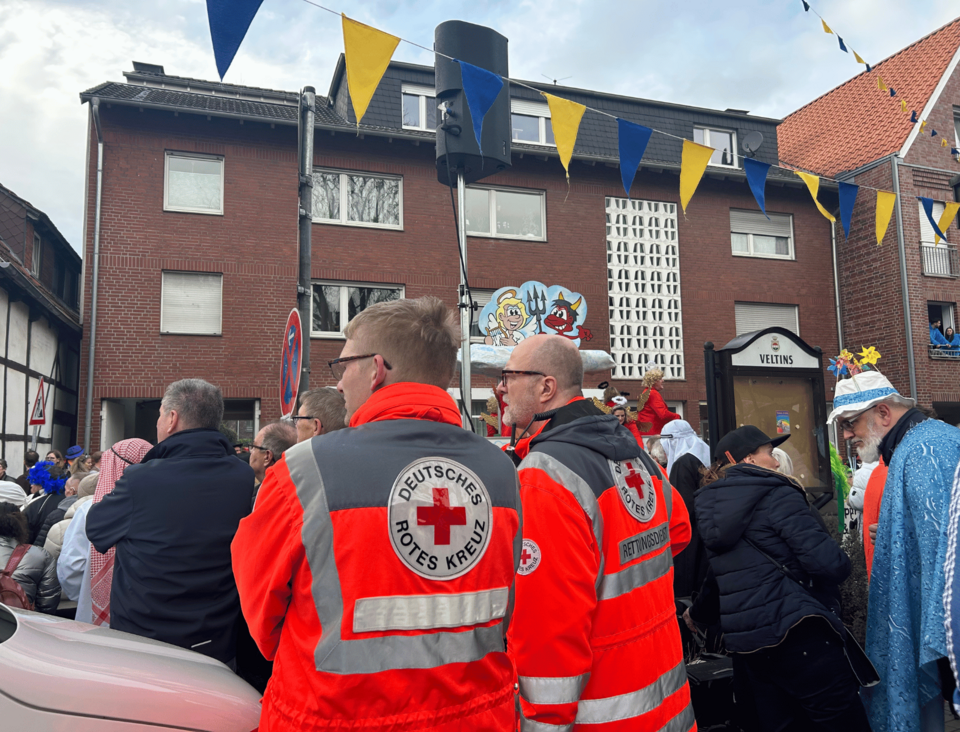Zwei Einsatzkräfte in Einsatzkleidung mit dem Rücken zur Kamera. Vor ihnen zieht der Karnevalszug durch Wolbeck.