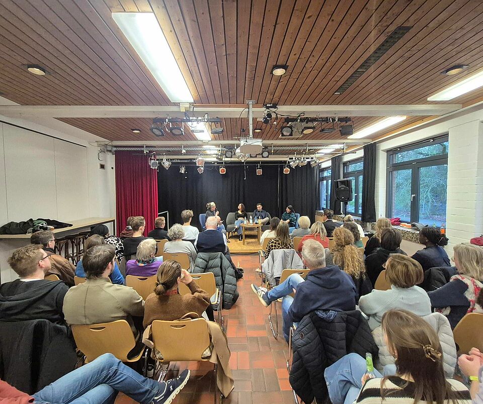 In einem großen Raum sitzen viele Menschen in Sitzreihen. Vor ihnen sitzen mehrere Personen auf einem Podium.