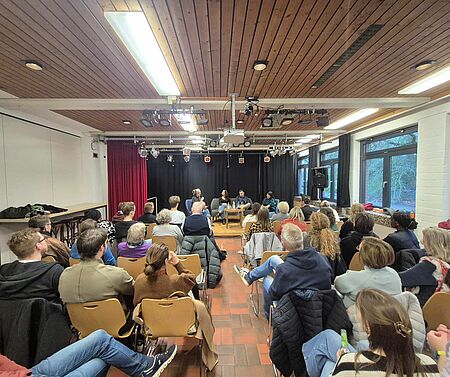 In einem großen Raum sitzen viele Menschen in Sitzreihen. Vor ihnen sitzen mehrere Personen auf einem Podium.
